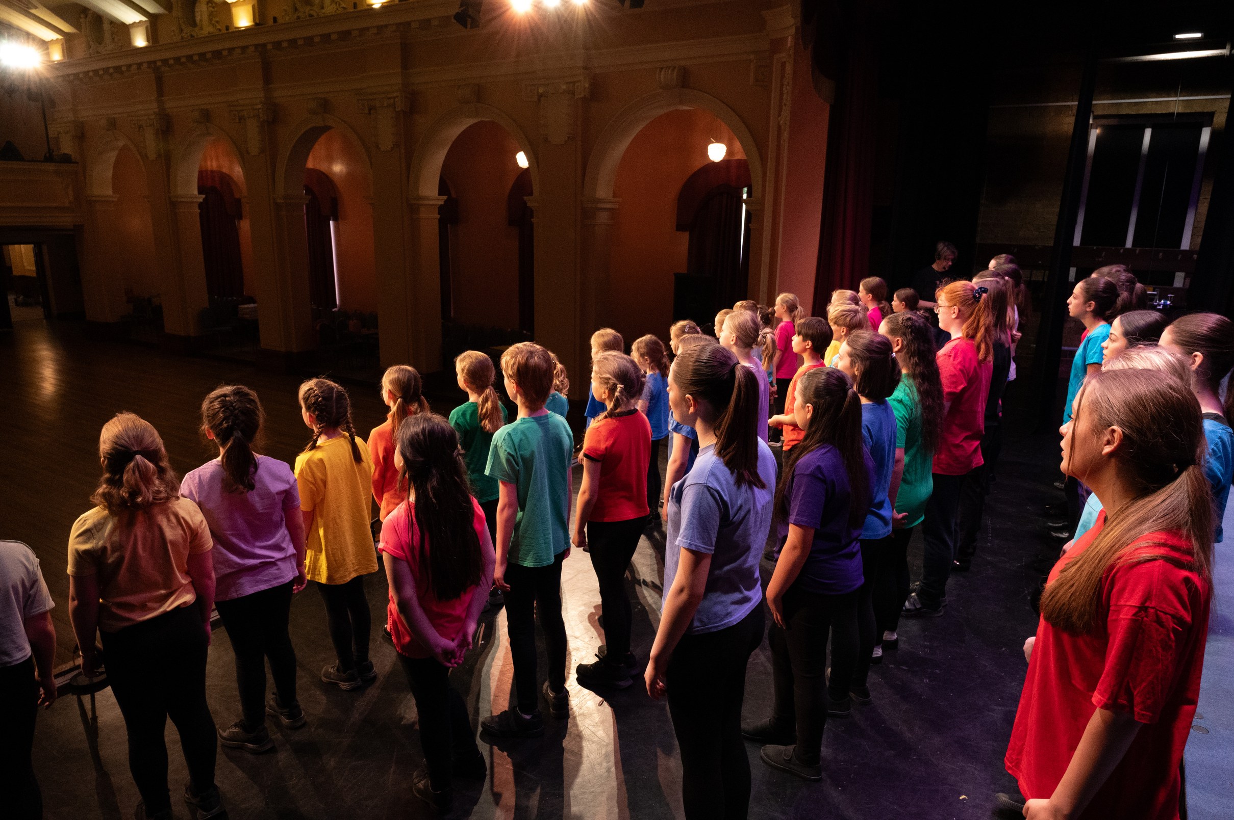 Cast singing together on stage in a grand hall