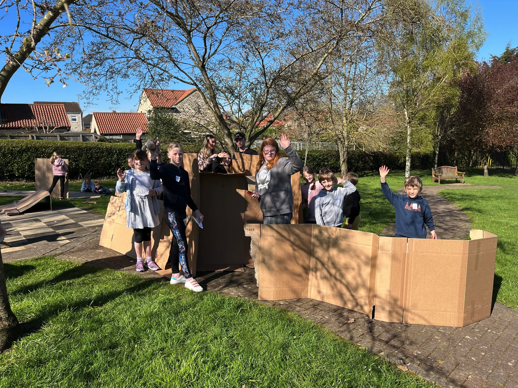 Children waving next to cardboard den walls in the park